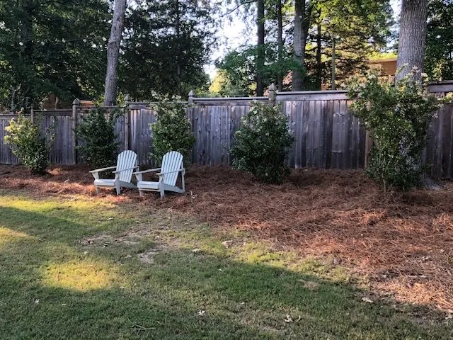 Backyard fence-line bed planted with shrubs and dressed in pine straw, with two white chairs, a Groundcare Landscape job
