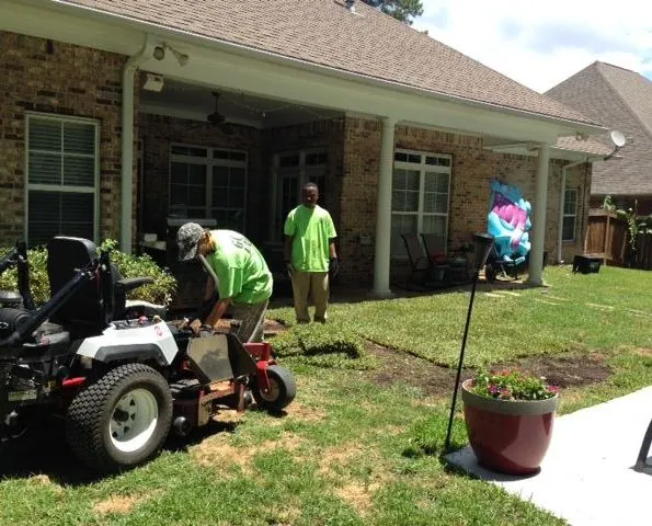 Groundcare Landscape crew in lime green shirts laying sod beside a zero-turn mower at a brick home